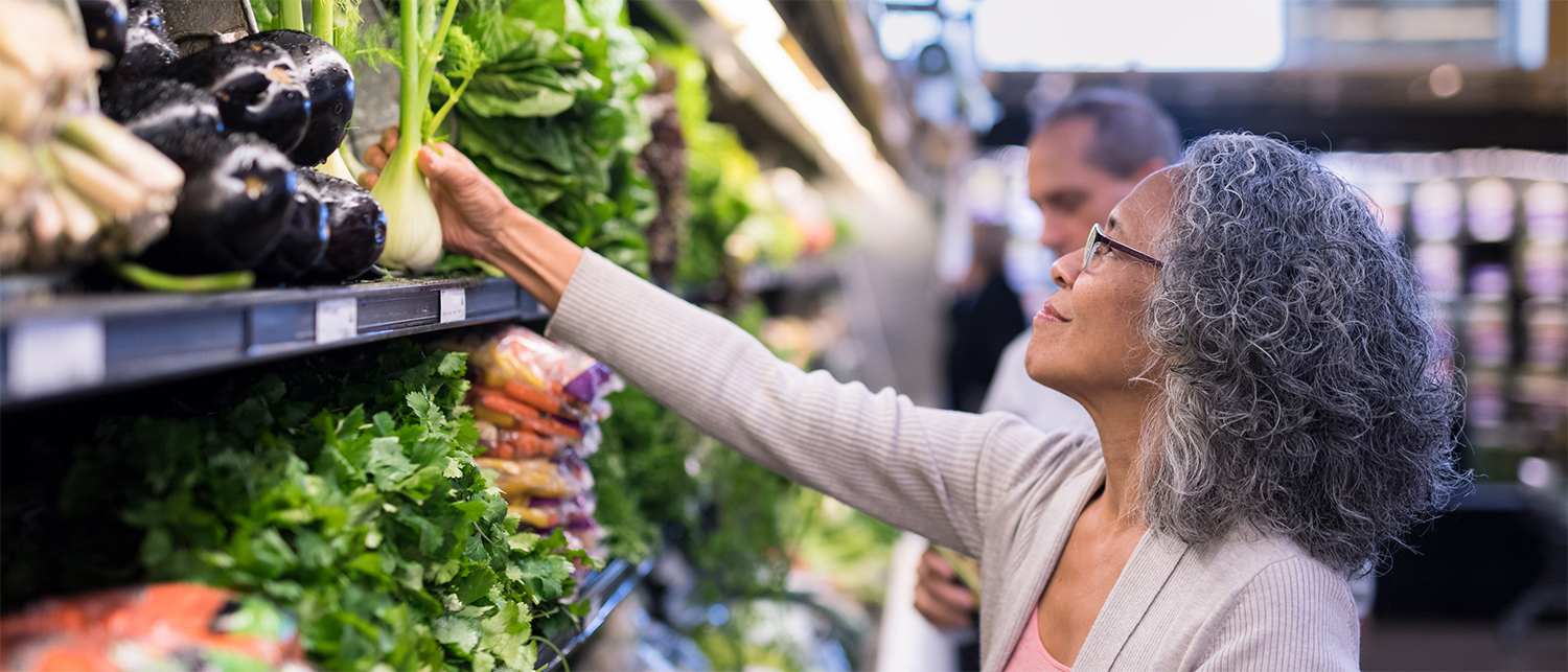 An older woman with glasses selects fresh fennel from a produce shelf in a grocery store, surrounded by leafy greens and vegetables.