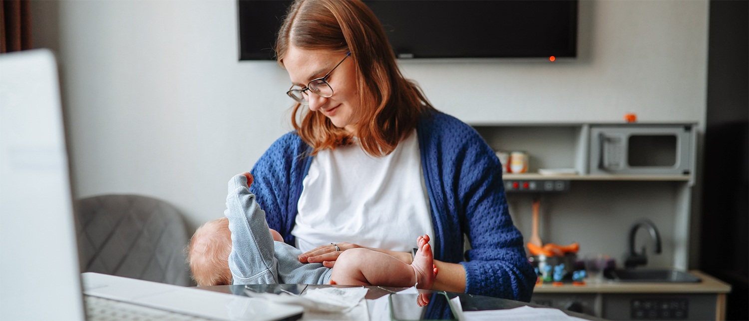 A woman wearing glasses looking at the baby on her lap while sitting at a desk with a laptop.