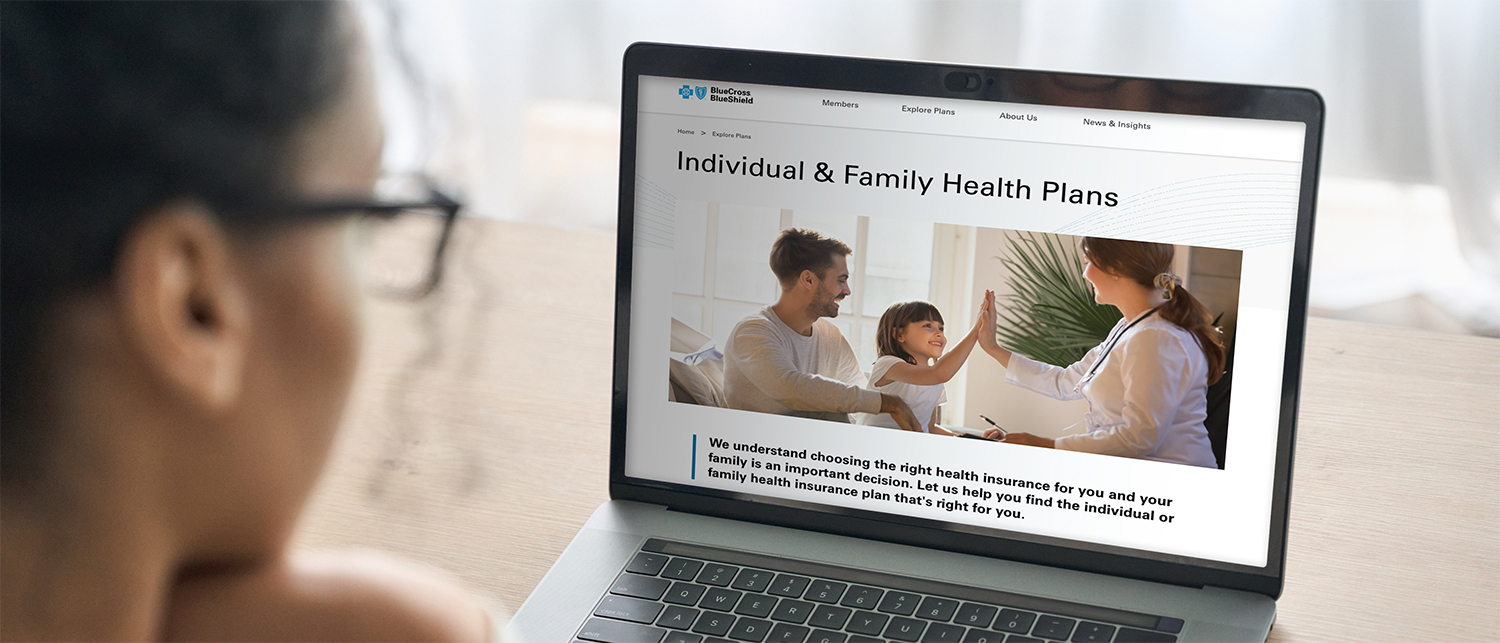 A person views a laptop screen displaying a health insurance website. The page highlights open enrollment, featuring a photo of a smiling family and a doctor, with text about individual and family health insurance plans.