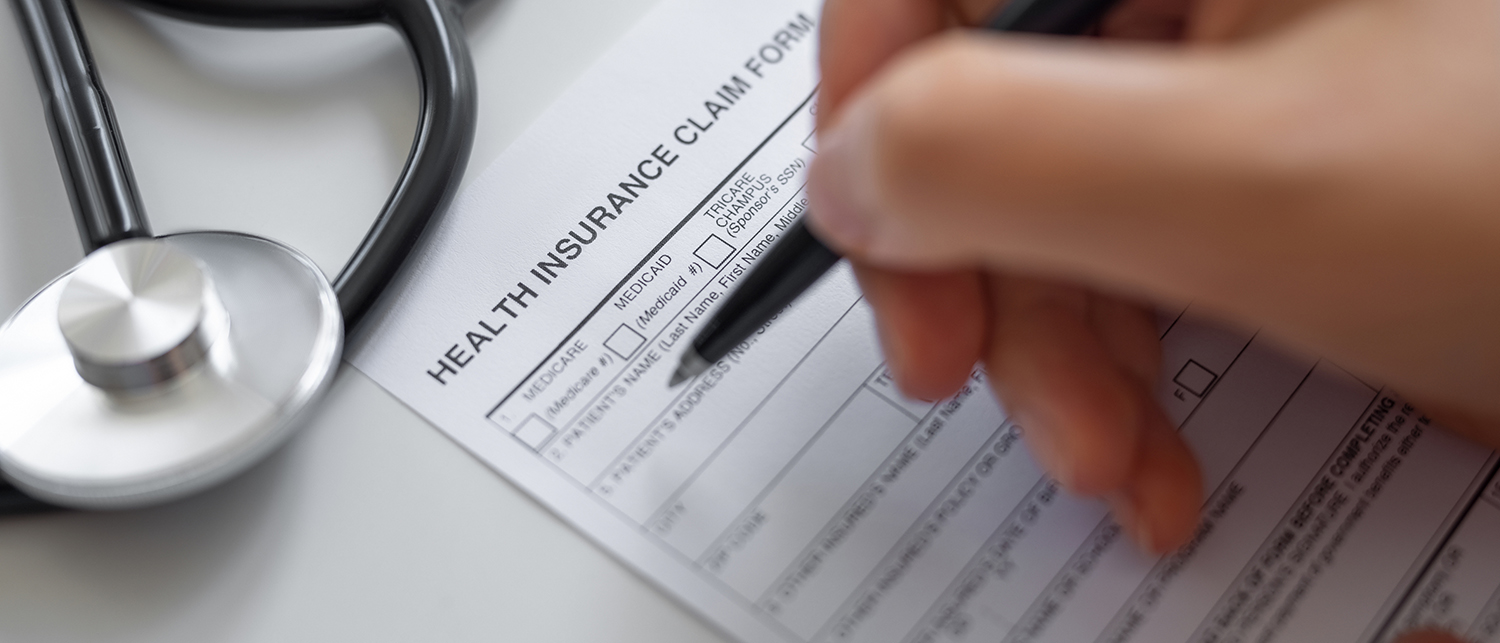 A person fills out a health insurance claim form with a pen. A stethoscope rests on the table beside the form.