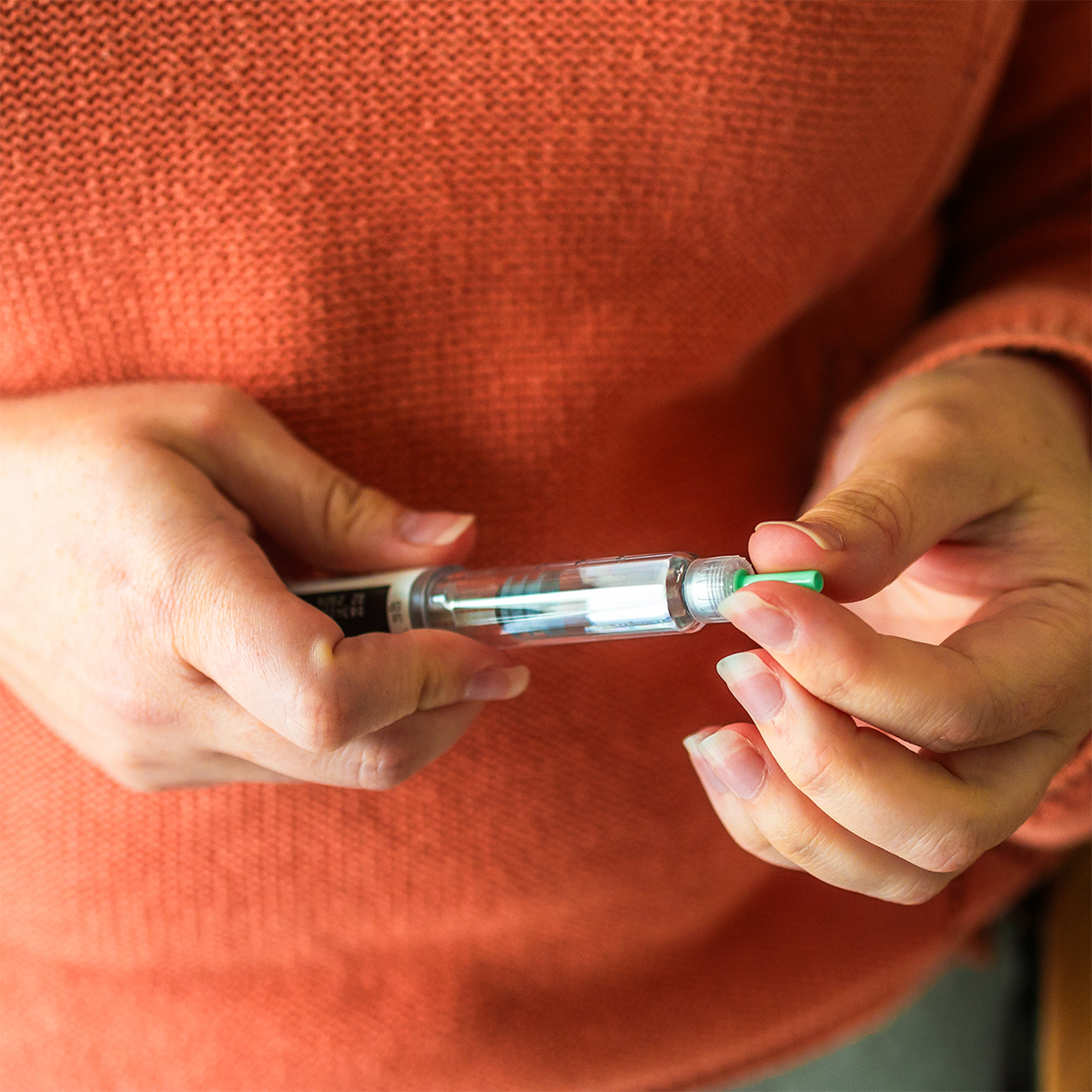 A person wearing an orange sweater holds an insulin pen, preparing to use it by removing the green cap to manage their insulin levels.