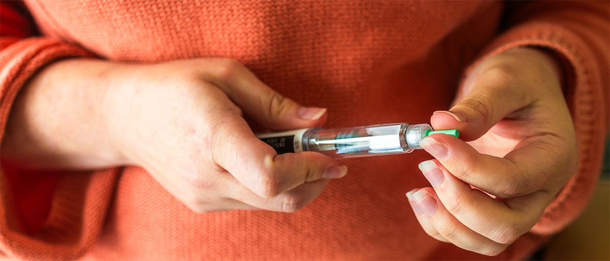 A person wearing an orange sweater holds an insulin pen, preparing to use it by removing the green cap to manage their insulin levels.